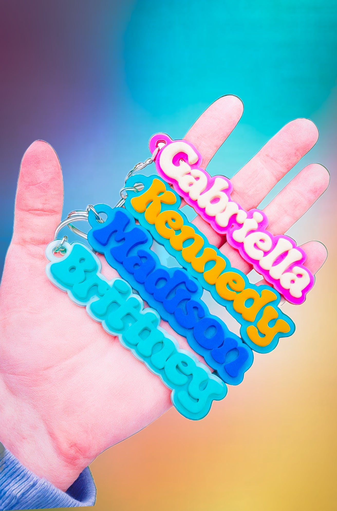 a hand holding four keychains with different names on them: Gabriella, Kennedy, Madison, and Brittaey. The keychains are in pastel colors and have a playful, rounded font style.